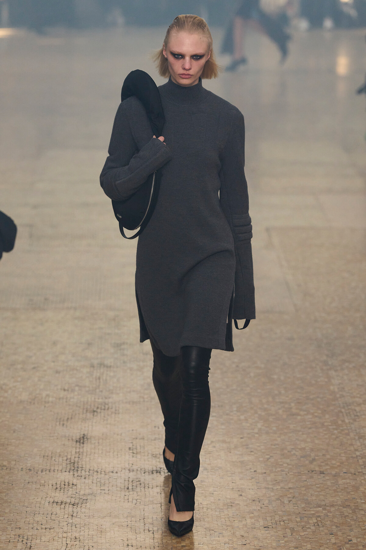Model walking on a runway in a dark gray turtleneck sweater dress, carrying a black circular bag over her shoulder with bold smoky eye makeup.