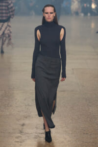 Model walking runway in a black sleeveless-turtleneck top with cutouts at the shoulders and a long gray skirt with side slits, black ankle boots.