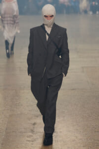 Runway model in a dark brown suit with a beige knit balaclava and a crossbody bag, walking forward on a fashion show runway.