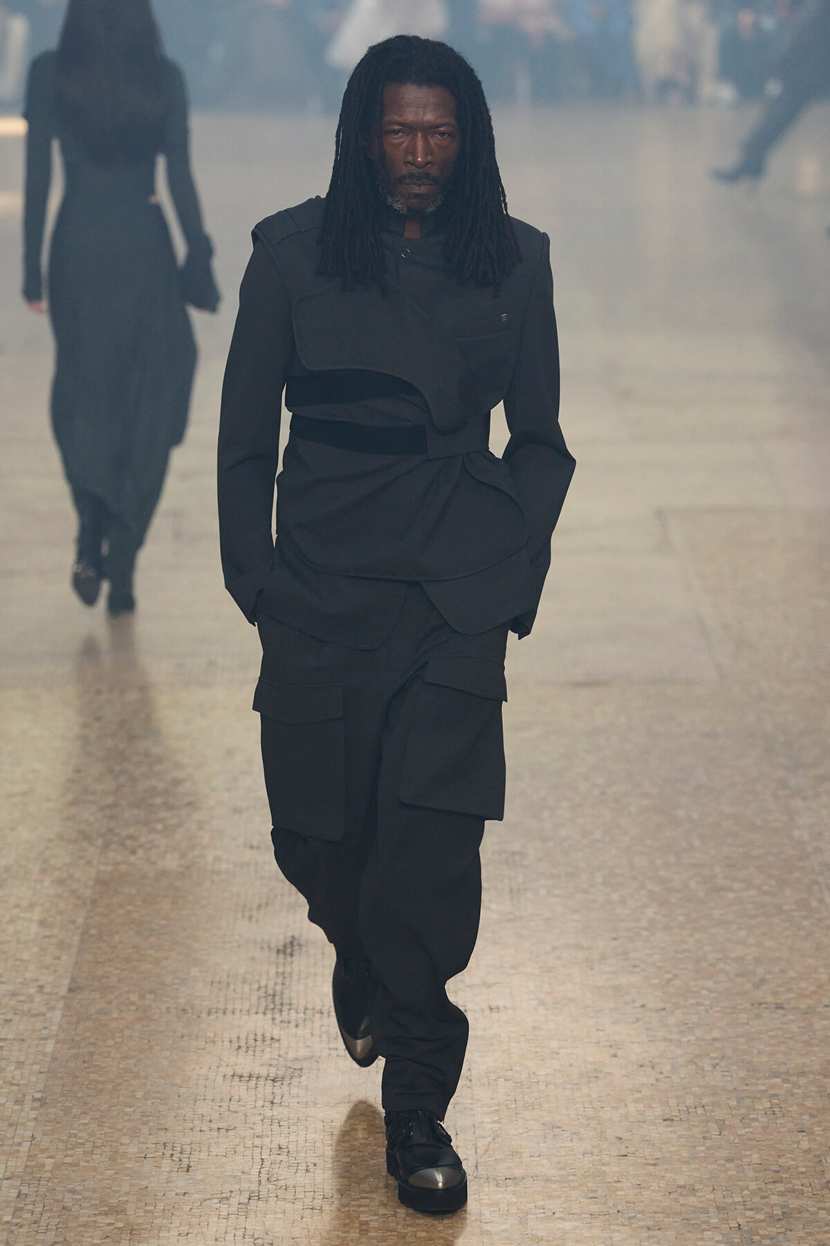 Model walking a fashion runway in an all-black, asymmetrical outfit with cargo pants and a belt, hands in pockets, dreadlocks flowing.