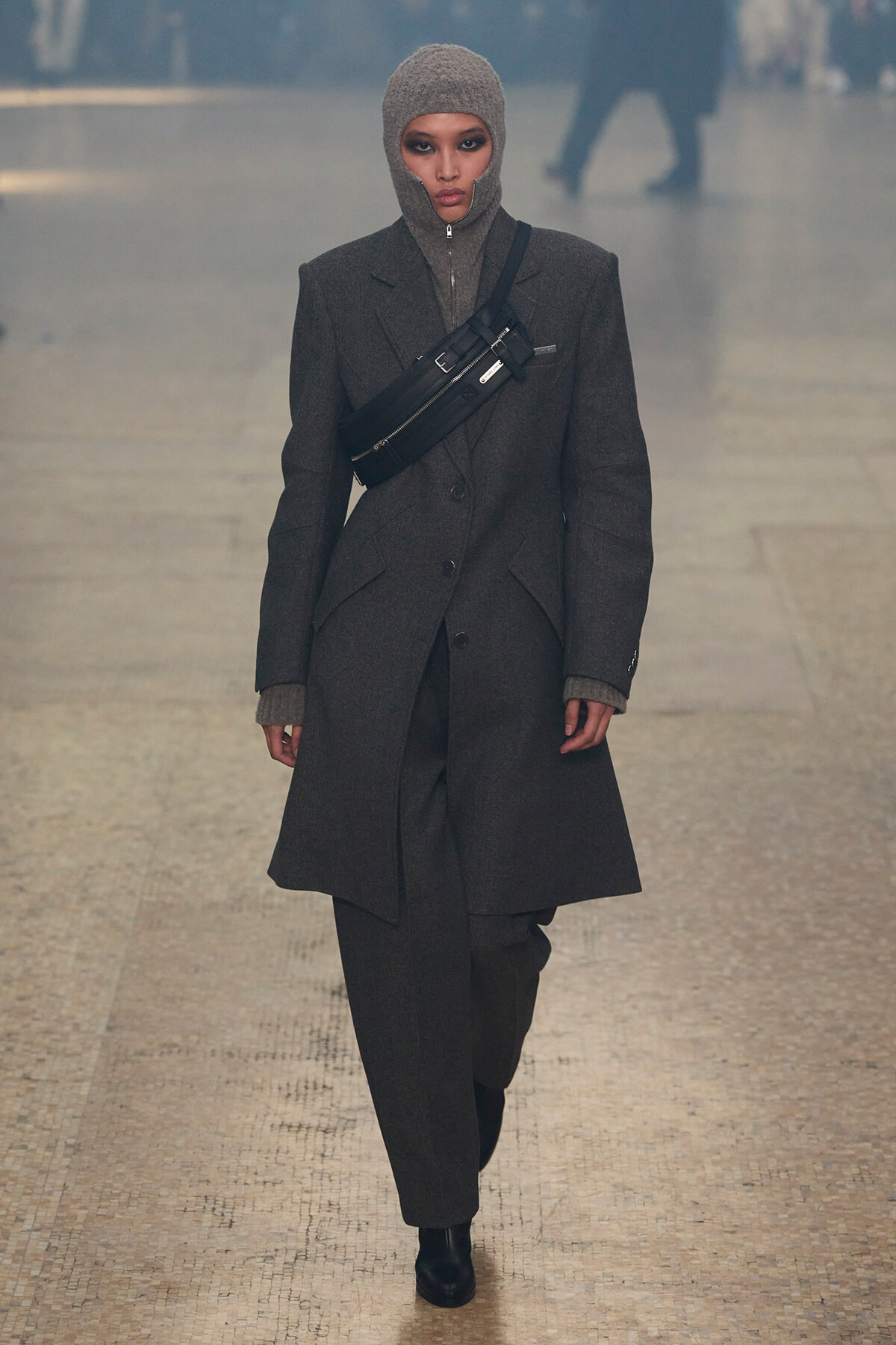 Model walking on a runway in a charcoal wool coat, matching knit balaclava, and a diagonal black crossbody bag across the chest.