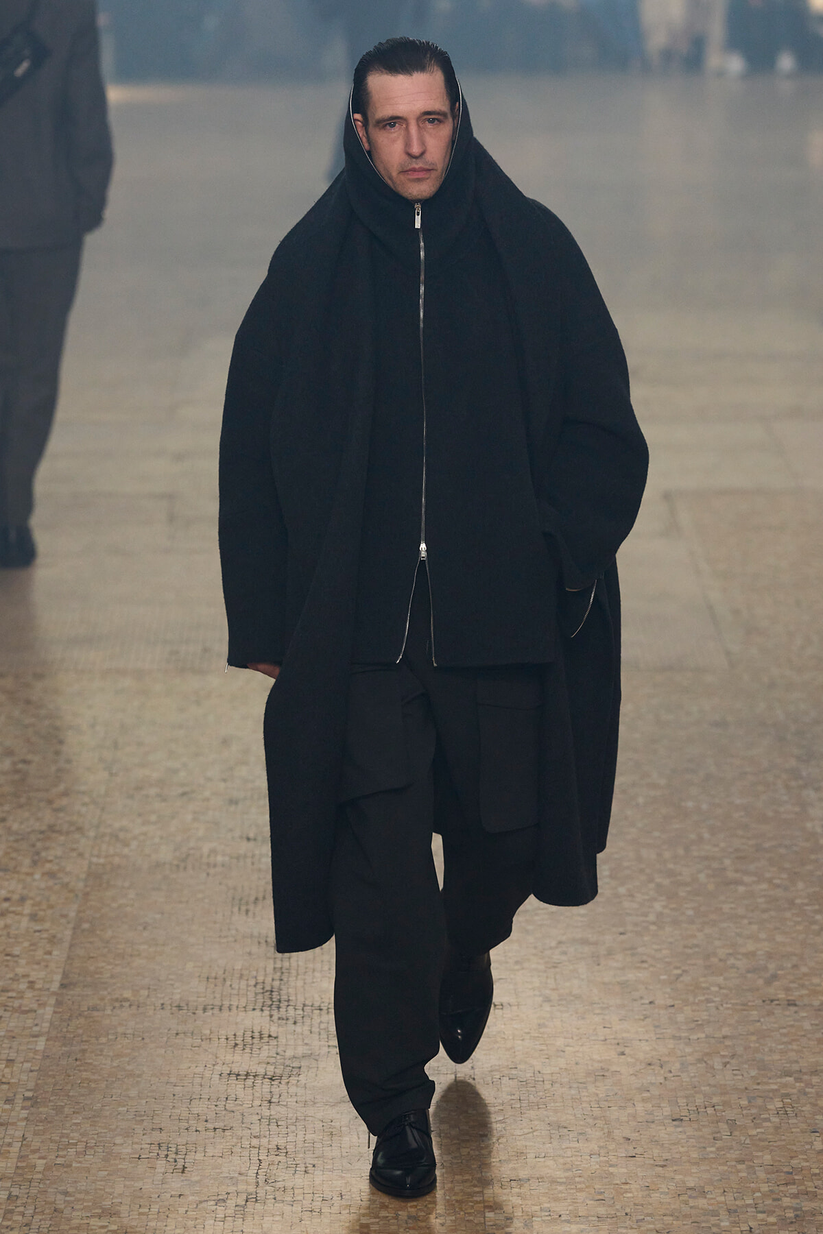 Man wearing an all-black oversized coat with the hood up, walking toward the camera in a tiled indoor corridor.