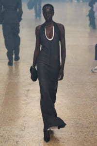 Model walking the runway in a dark sleeveless halter gown with a white-trimmed neckline, carrying a small black clutch on a tiled catwalk in dim lighting.