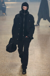 Person dressed in all black, wearing a hooded balaclava and zip-front jacket, walking toward the camera in a pale tiled indoor hallway while carrying a black bag