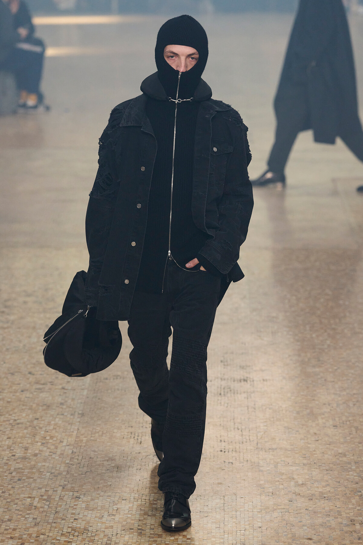 Person dressed in all black, wearing a hooded balaclava and zip-front jacket, walking toward the camera in a pale tiled indoor hallway while carrying a black bag