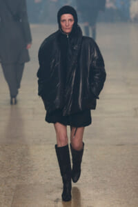 Person walking toward the camera in a glossy black oversized puffer jacket with hood, black dress, and knee‑high boots in a foggy indoor walkway.