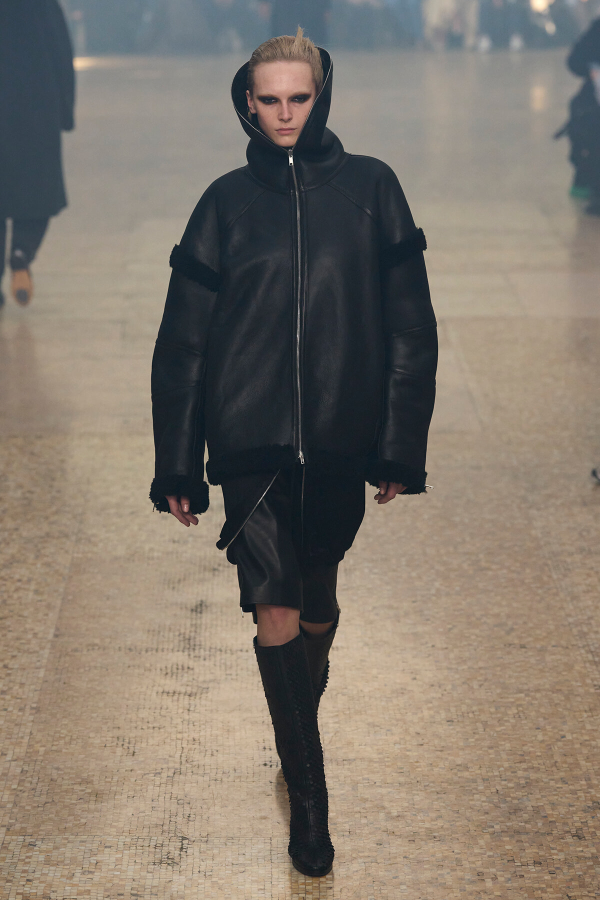 Model walks the runway in an oversized black leather zip-up jacket with a large hood, paired with tall black boots on a neutral runway.