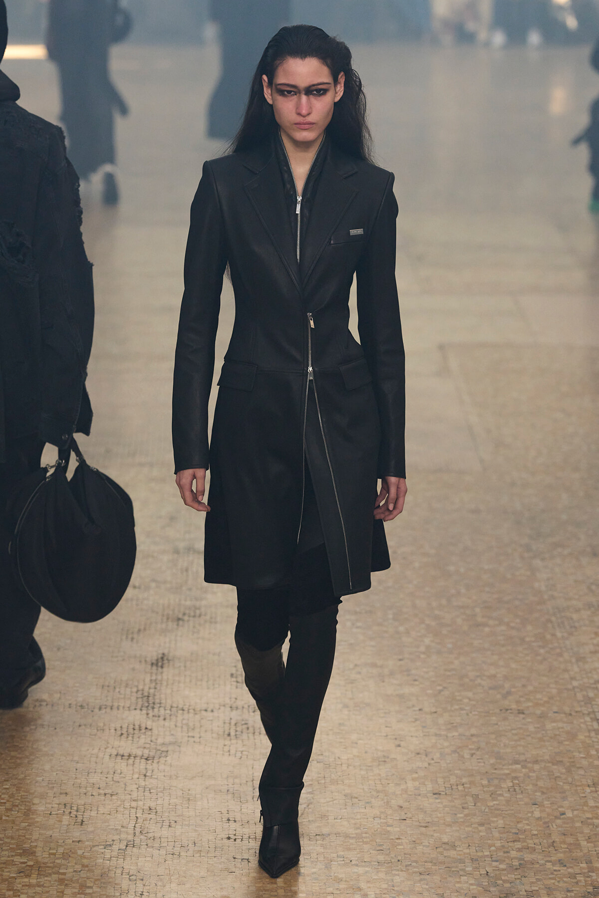 Female model walks a runway in a long black tailored coat with zippers, wearing black boots and a serious expression.