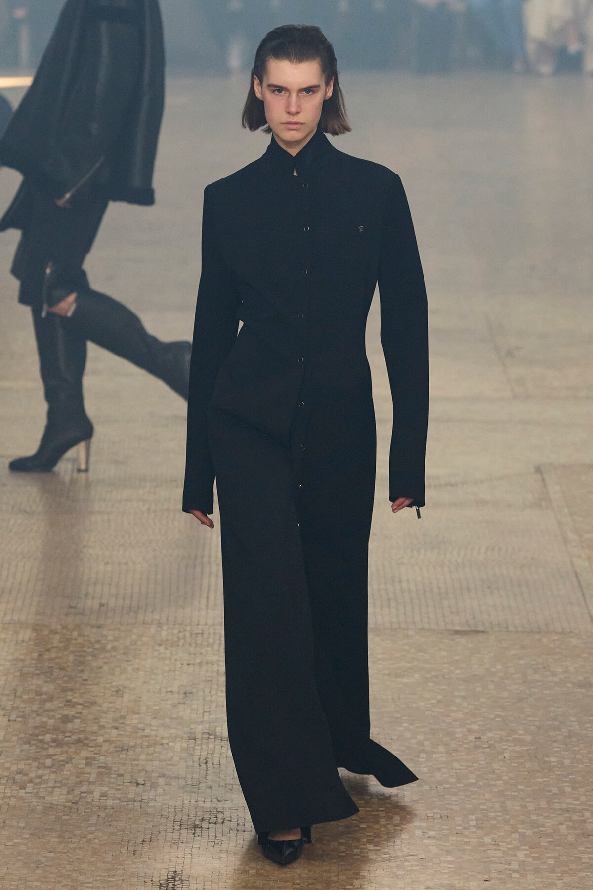 Model walking a runway in a long black tailored coat and wide-leg pants on a tiled catwalk.