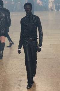 Model walks the runway in an all-black, studded knit top and matching pants with a belt on a foggy catwalk.