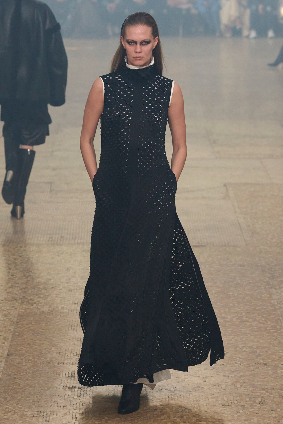 Fashion model walking the runway in a sleeveless black, textured gown with cutout details and a white collar, stern expression.