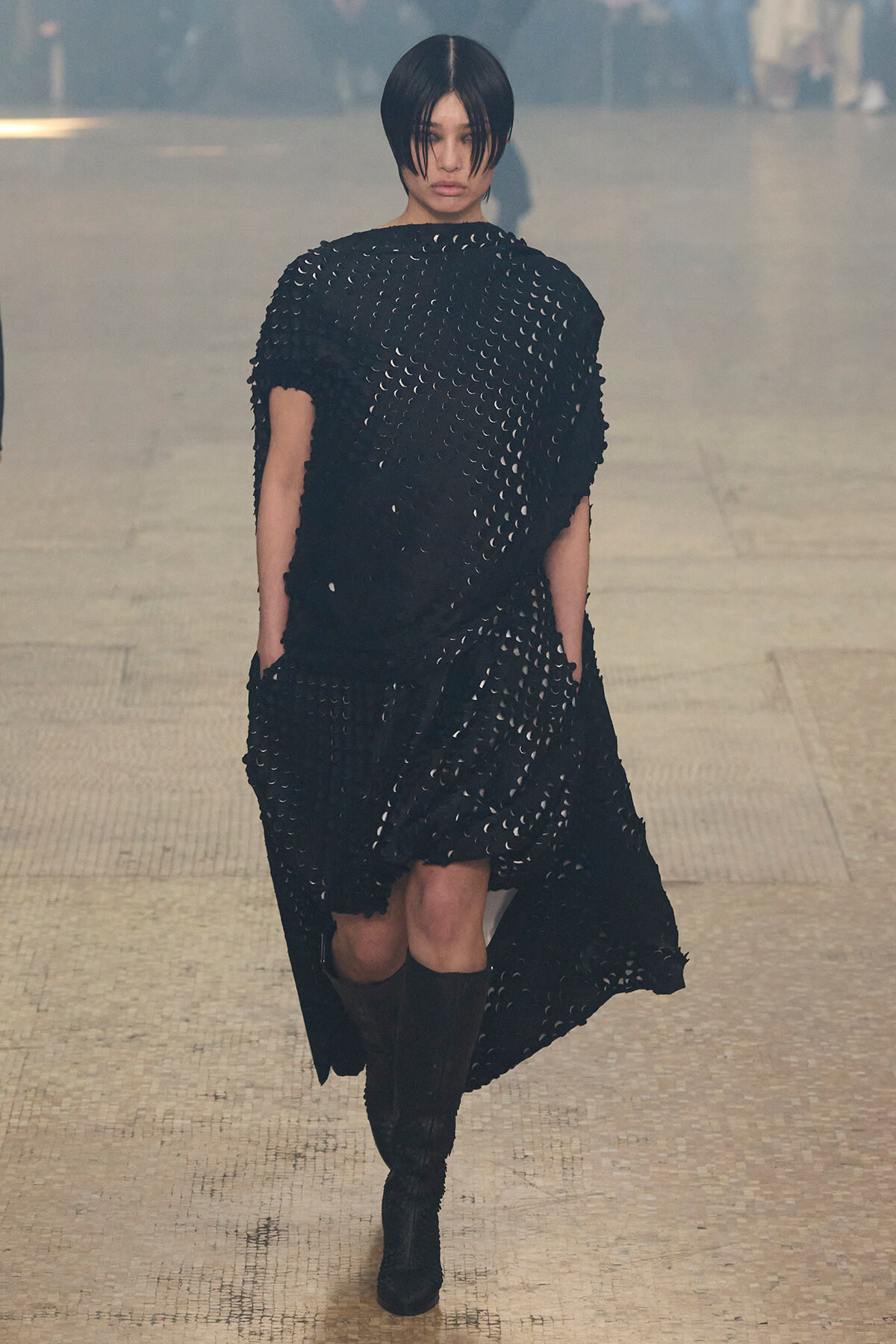 Model on a runway wearing a black dress with circular cutouts and knee-high boots, walking forward.