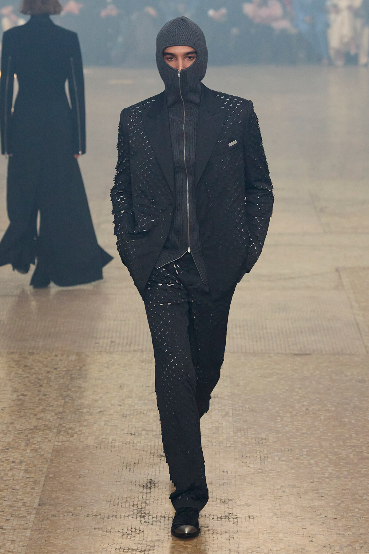 Model walking a fashion runway in a black studded blazer and trousers, wearing a zip-up balaclava hood for a futuristic look.