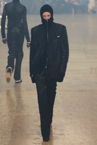 Model walks the runway in a black, studded suit with a balaclava hood and zipper front against a smoky backdrop.