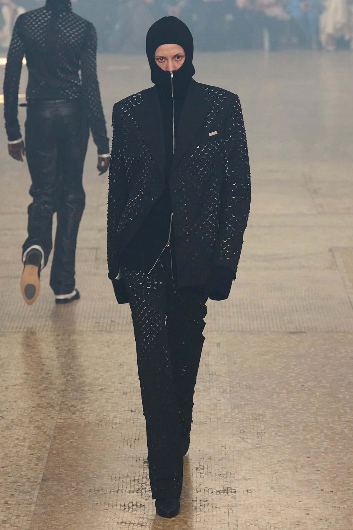 Model walks the runway in a black, studded suit with a balaclava hood and zipper front against a smoky backdrop.