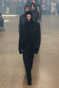 Model walks a fashion show wearing a black hooded coat with studded details, followed by others in matching outfits as an audience watches from the background.