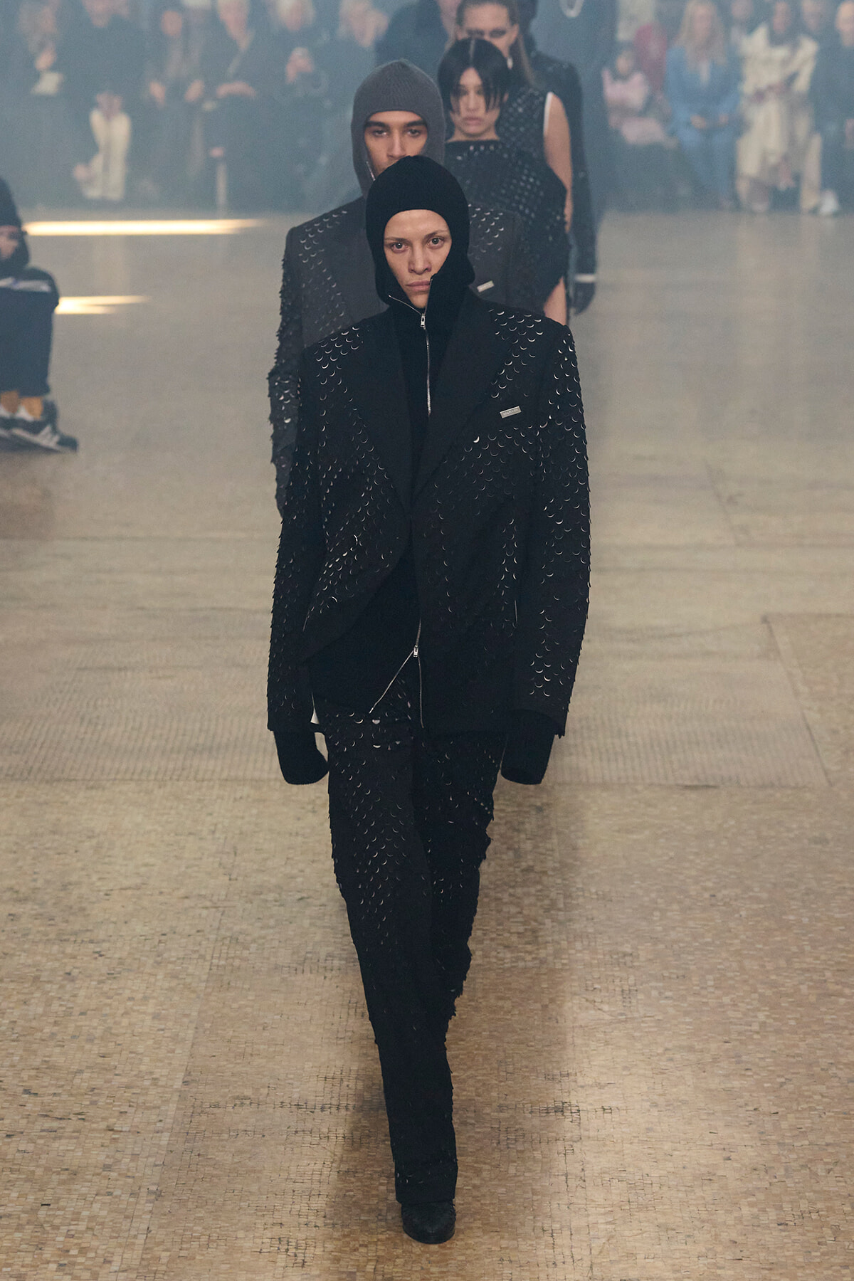 Model walks a fashion show wearing a black hooded coat with studded details, followed by others in matching outfits as an audience watches from the background.