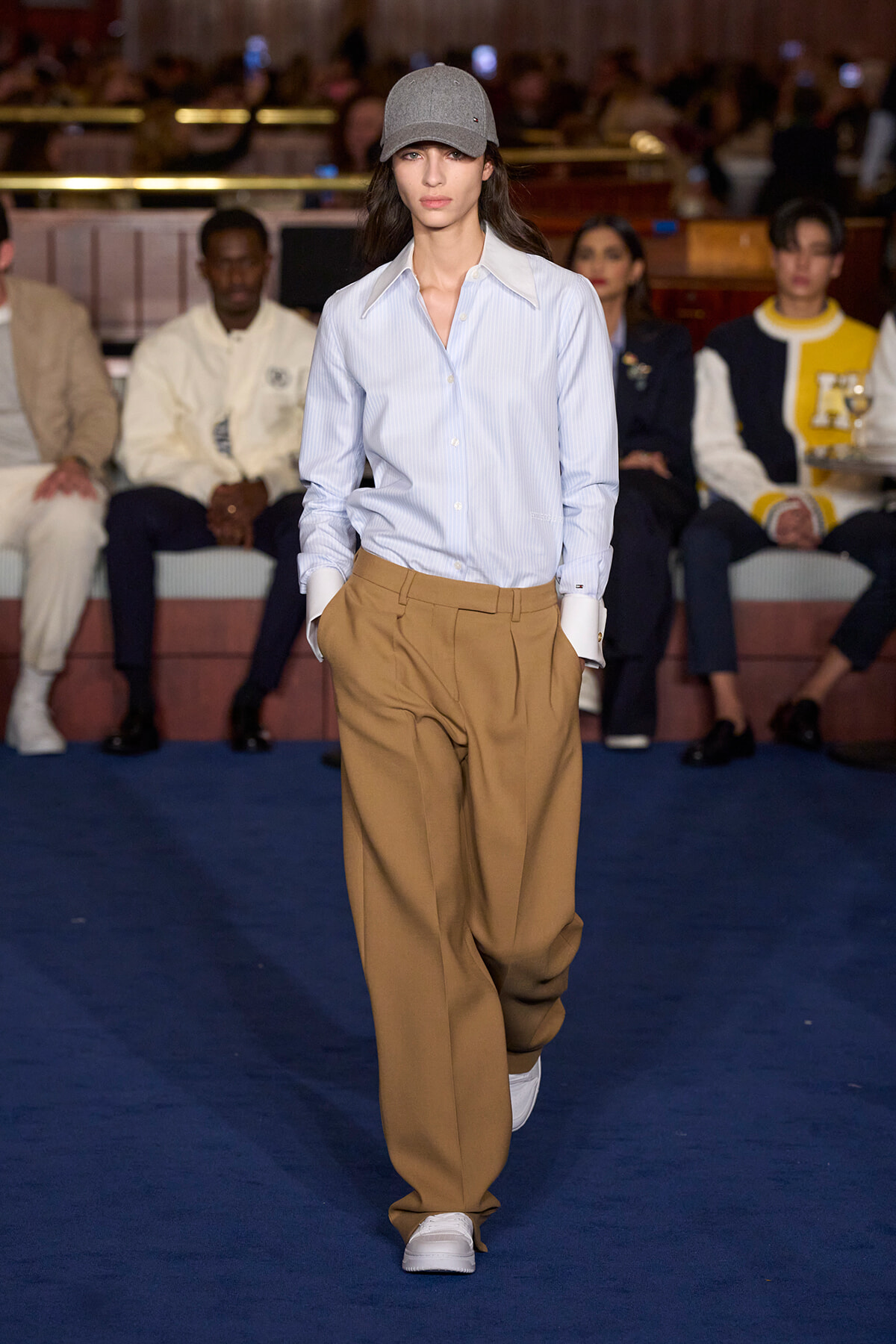 Model walking a runway in a light blue striped shirt, tan wide-leg trousers, white sneakers, and a gray cap, with an audience in the background.