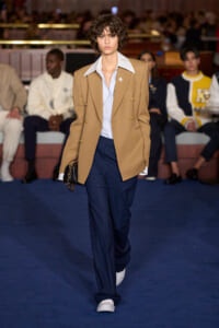 Model walking a runway in a tan double‑breasted blazer, light blue shirt, navy trousers, and white sneakers, carrying a small black bag.
