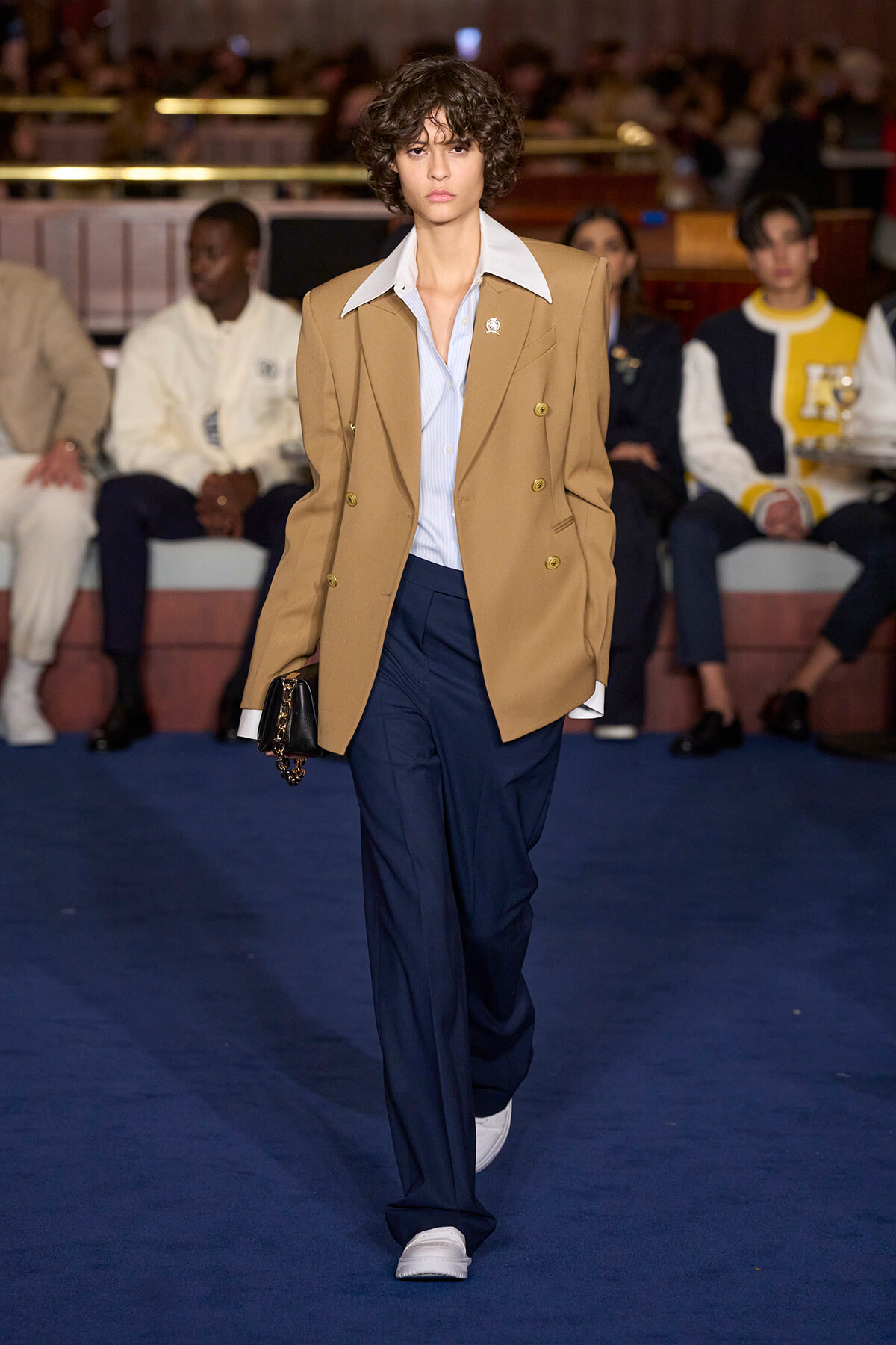Model walking a runway in a tan double‑breasted blazer, light blue shirt, navy trousers, and white sneakers, carrying a small black bag.