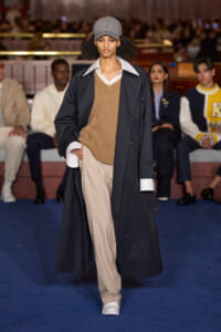 Runway model walking the catwalk in a long navy trench over a tan sweater and beige trousers, with a gray cap and white sneakers; audience in background.