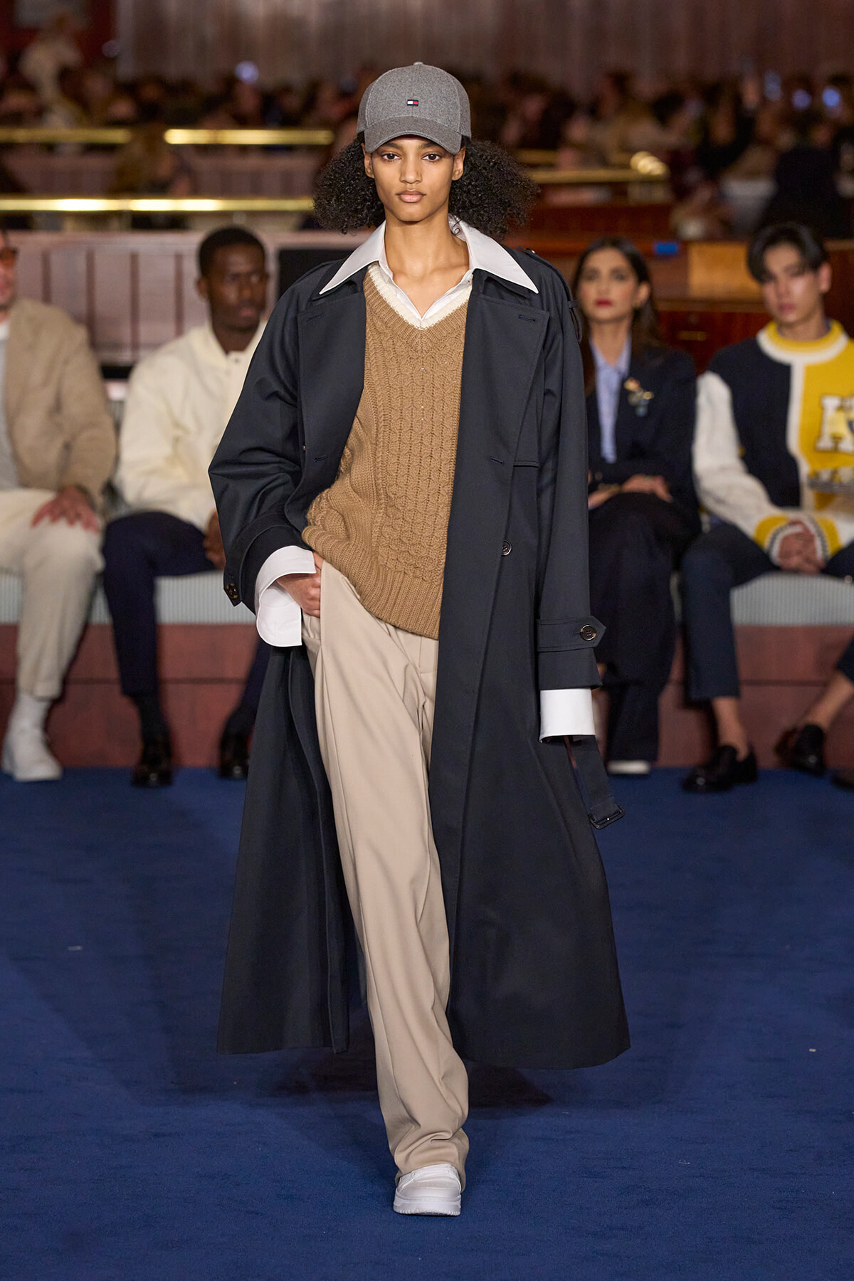 Runway model walking the catwalk in a long navy trench over a tan sweater and beige trousers, with a gray cap and white sneakers; audience in background.