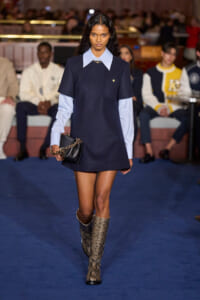Model walking a fashion runway in a navy shift dress with a pale blue oversized collar, carrying a black bag with a gold chain.