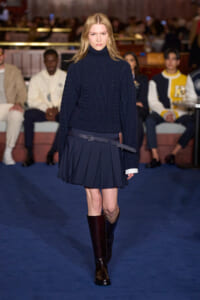 Model on a runway wearing a navy cable-knit turtleneck sweater with a matching pleated navy skirt and belt, audience in the background.
