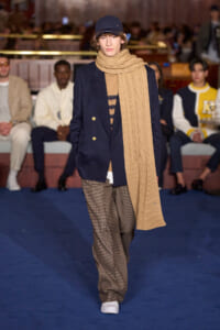 Model walking a runway wearing a navy blazer, tan knit scarf, and brown checkered trousers with white sneakers.