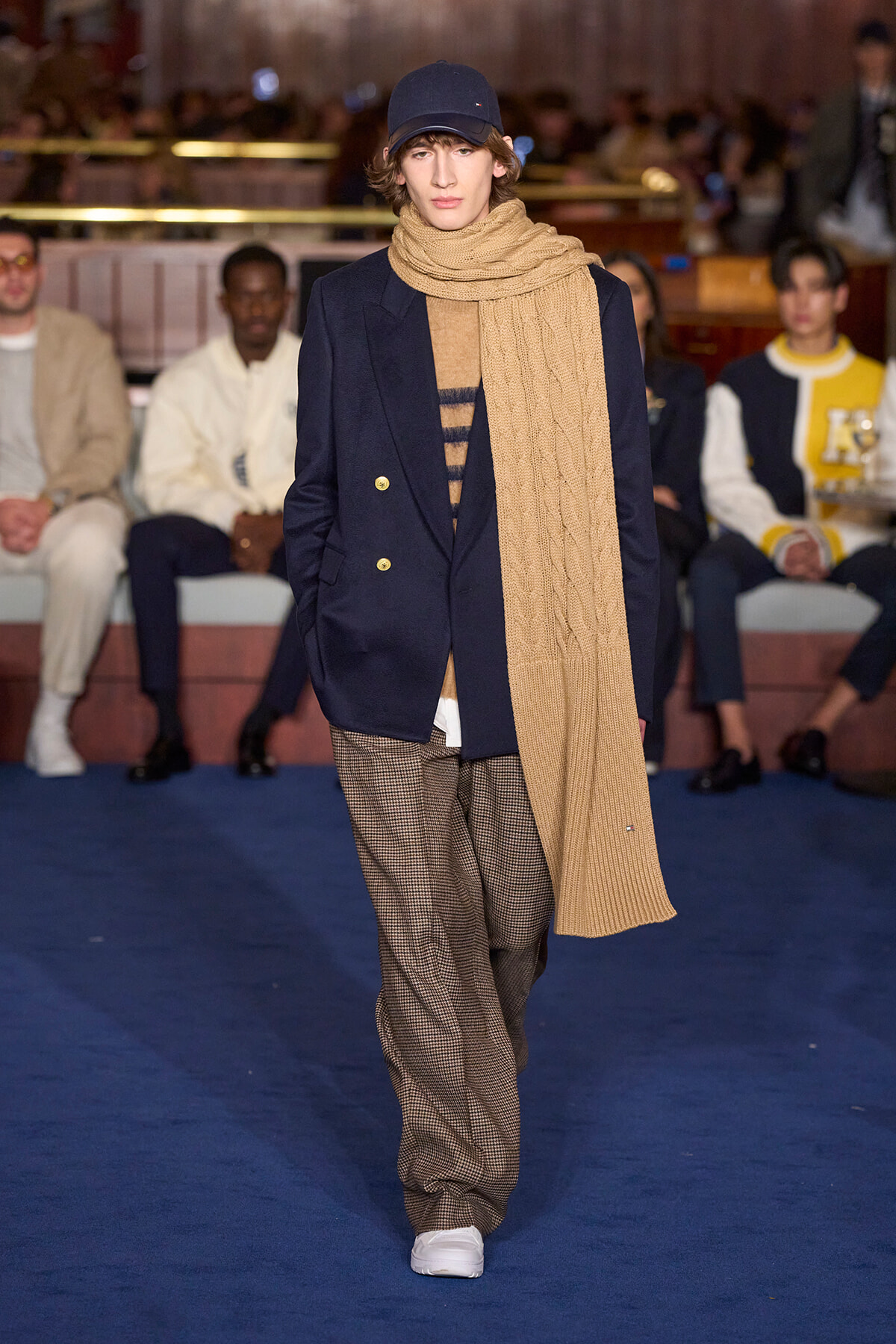 Model walking a runway wearing a navy blazer, tan knit scarf, and brown checkered trousers with white sneakers.