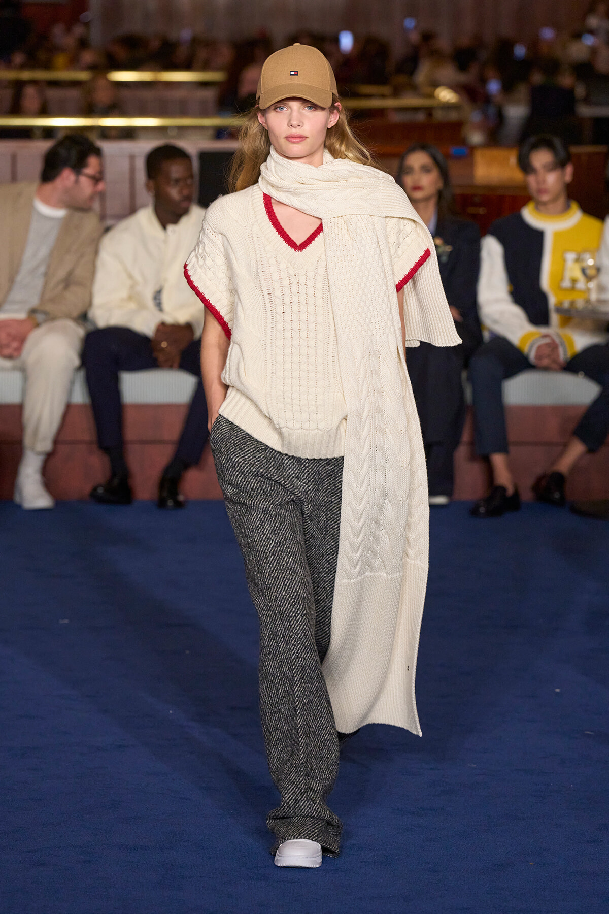 Model walking a runway in a cream knit sweater with red trim, oversized scarf, gray textured pants, and white sneakers; beige cap in indoor show.