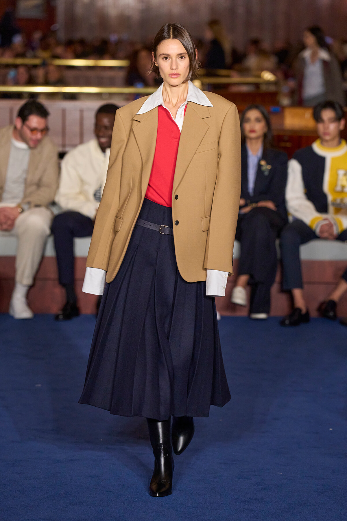 Model walking a runway in a camel blazer over a red top and white shirt, navy pleated skirt and black boots; audience blurred in background.