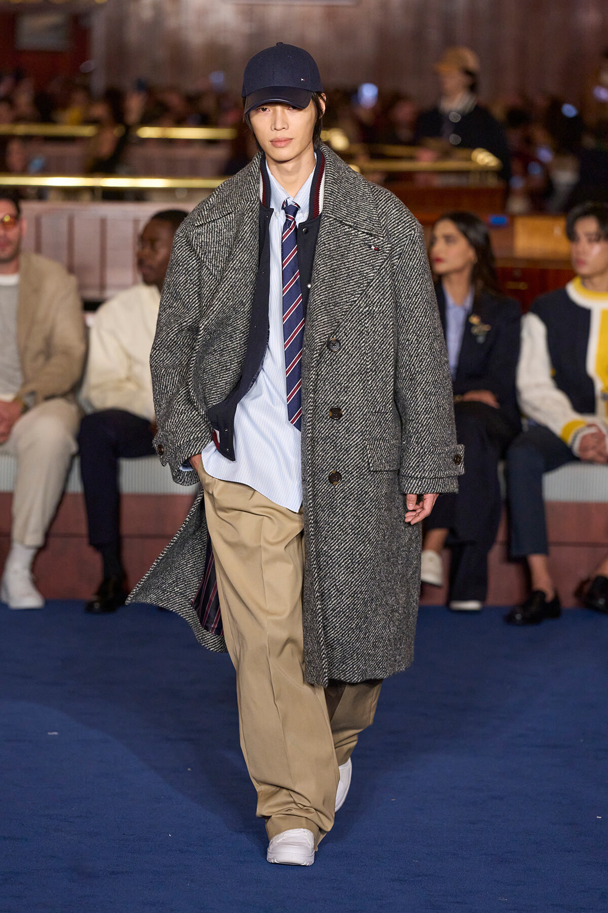 Model walking a runway in an oversized grey herringbone coat over a light blue shirt and striped tie; audience blurred in background.