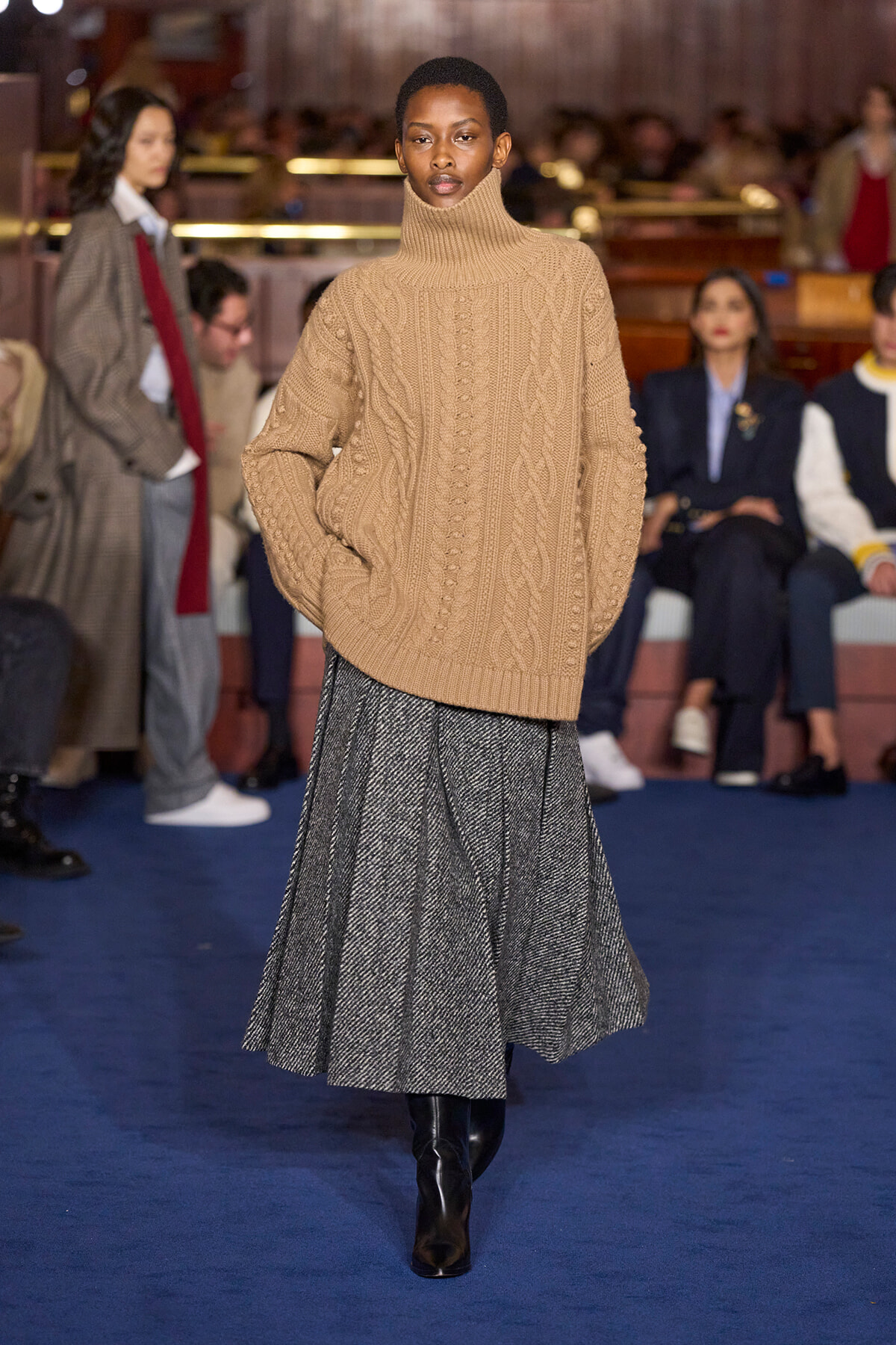 Model walks a runway in a chunky beige turtleneck sweater and gray pleated midi skirt with black boots; audience sits in the background.