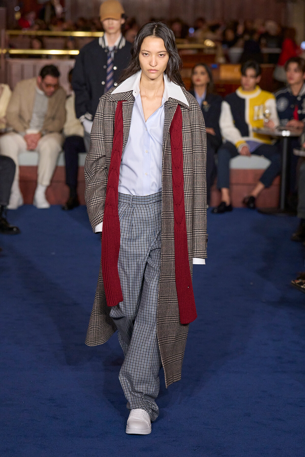 Model walking a fashion show runway in a long plaid coat with a red scarf, light blue shirt, and checked trousers.