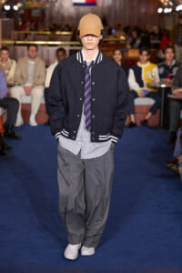 Male model walks a runway in a navy varsity jacket, striped shirt and tie, gray trousers, white sneakers, and a tan cap.