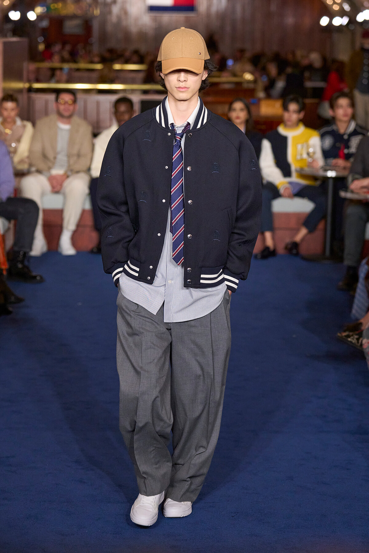 Male model walks a runway in a navy varsity jacket, striped shirt and tie, gray trousers, white sneakers, and a tan cap.