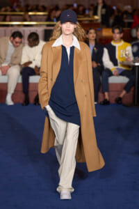 Model walks a runway in a camel overcoat over a navy shirt and white collared shirt, with light trousers and white sneakers. Cap worn.