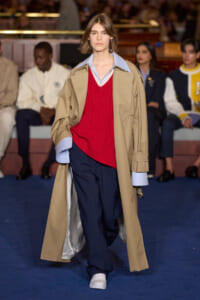 Model walks the runway in a tan trench coat over a red knit sweater and light blue shirt, paired with navy trousers and white sneakers.