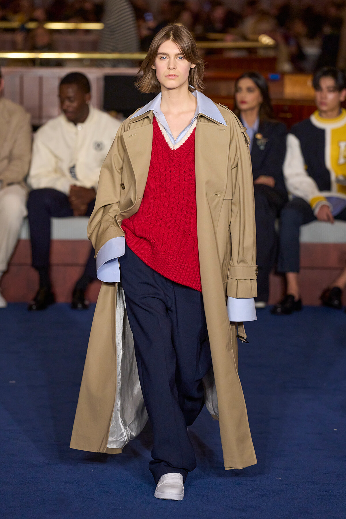 Model walks the runway in a tan trench coat over a red knit sweater and light blue shirt, paired with navy trousers and white sneakers.