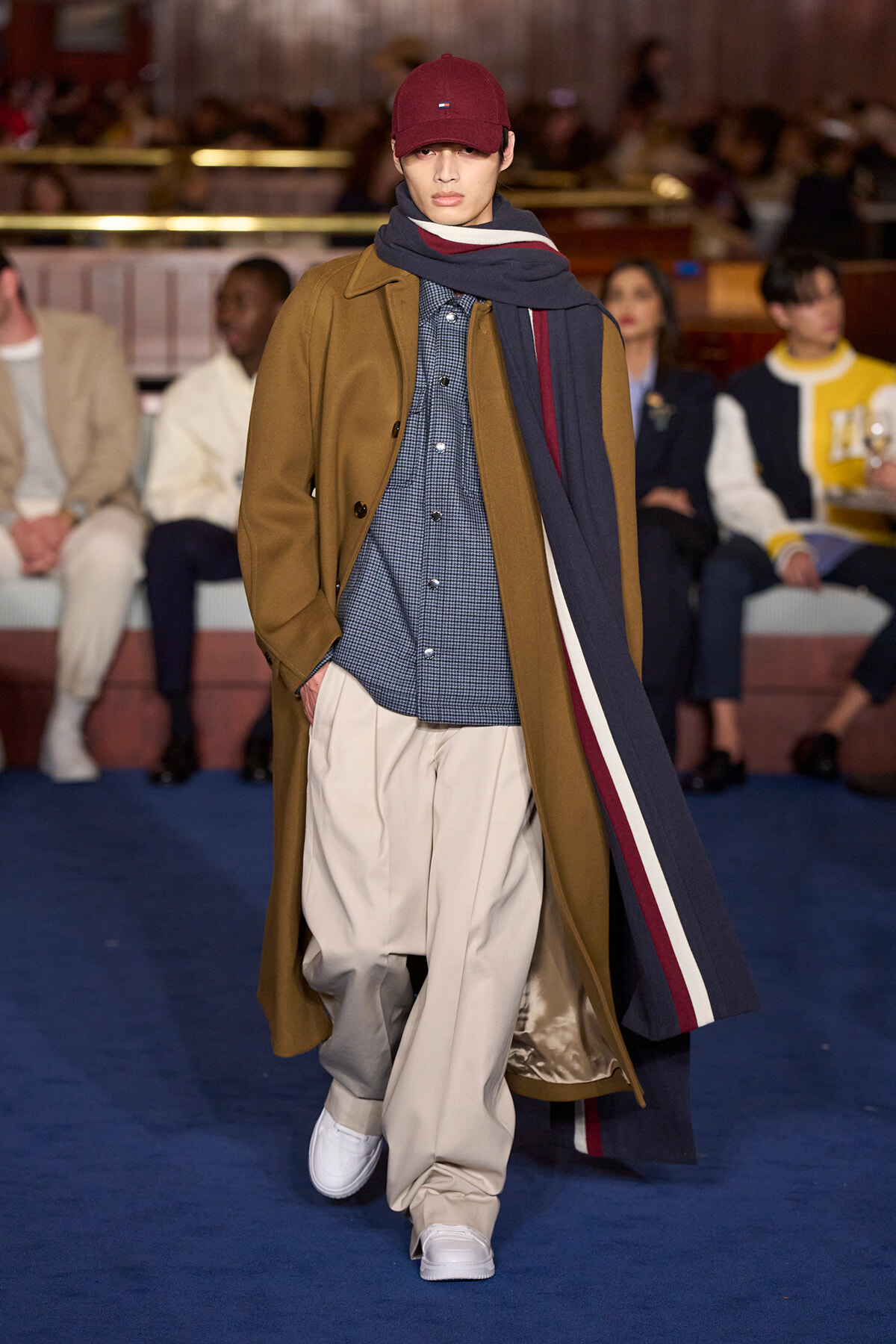 Model walking a fashion runway in a camel coat, checkered shirt, beige trousers, white sneakers, navy scarf, and red cap.