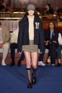 Model walks a fashion runway wearing a navy shearling-lined jacket over a cream knit sweater and houndstooth mini skirt with black knee-high socks and loafers, cap on head.