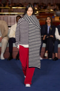 Model walking the runway in an oversized black-and-white striped knit coat draped over red pants, white sneakers, with a blue carpet and seated audience in the background.