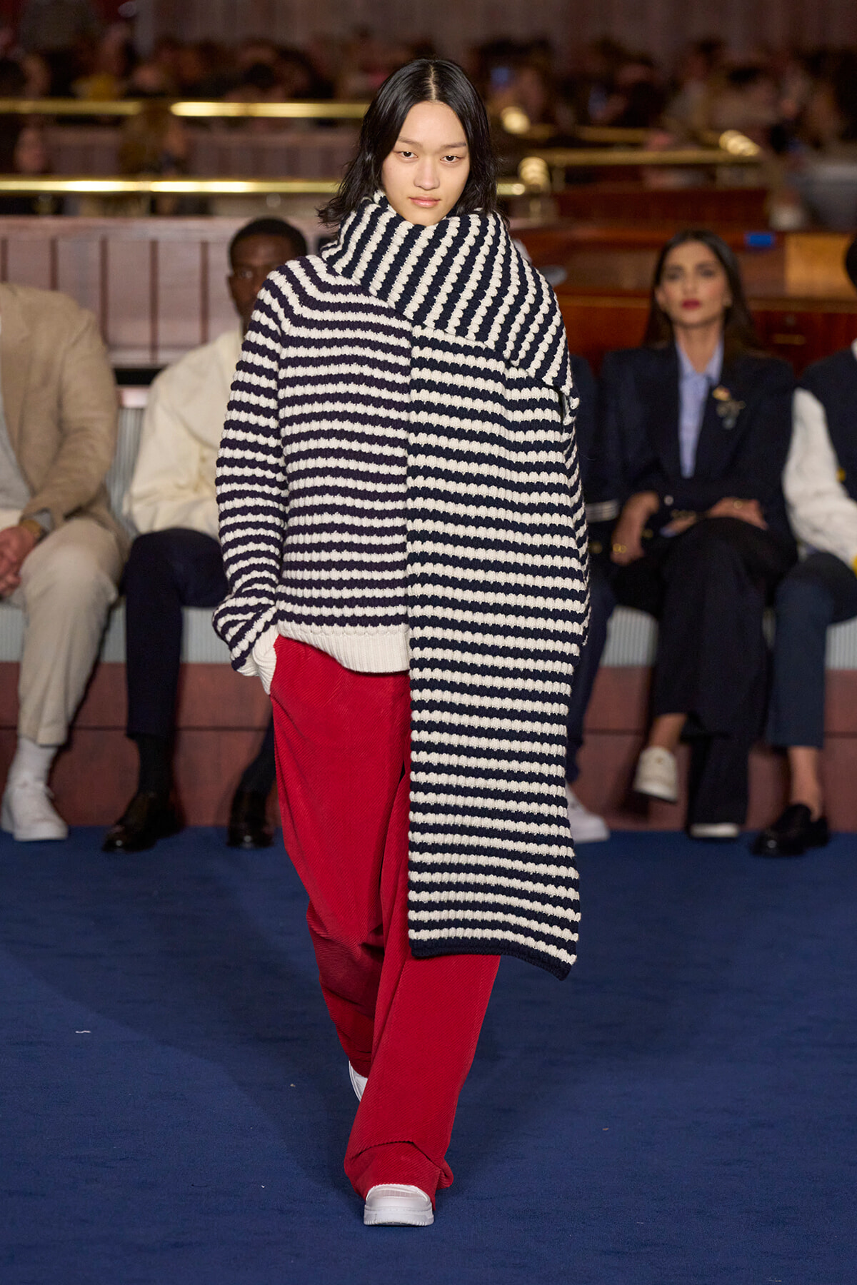 Model walking the runway in an oversized black-and-white striped knit coat draped over red pants, white sneakers, with a blue carpet and seated audience in the background.