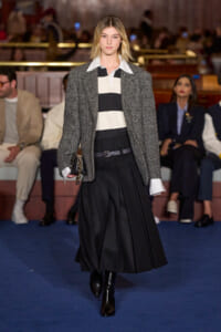 Runway model walks the catwalk in a gray herringbone blazer over a black-and-white striped top and a long black pleated skirt, clutch in hand.