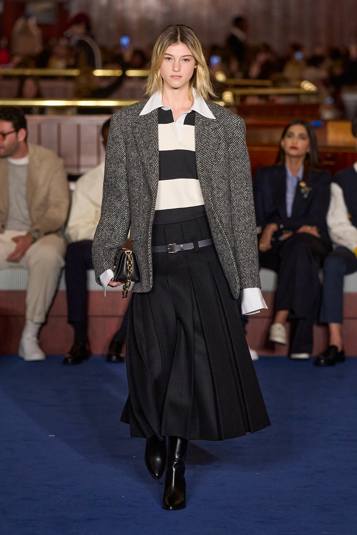 Runway model walks the catwalk in a gray herringbone blazer over a black-and-white striped top and a long black pleated skirt, clutch in hand.