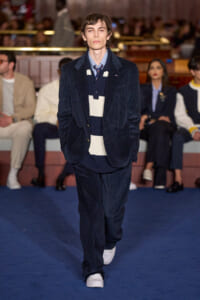 Male model walks down runway in navy corduroy suit with striped navy-and-cream sweater and tie at a fashion show. Audience in background.