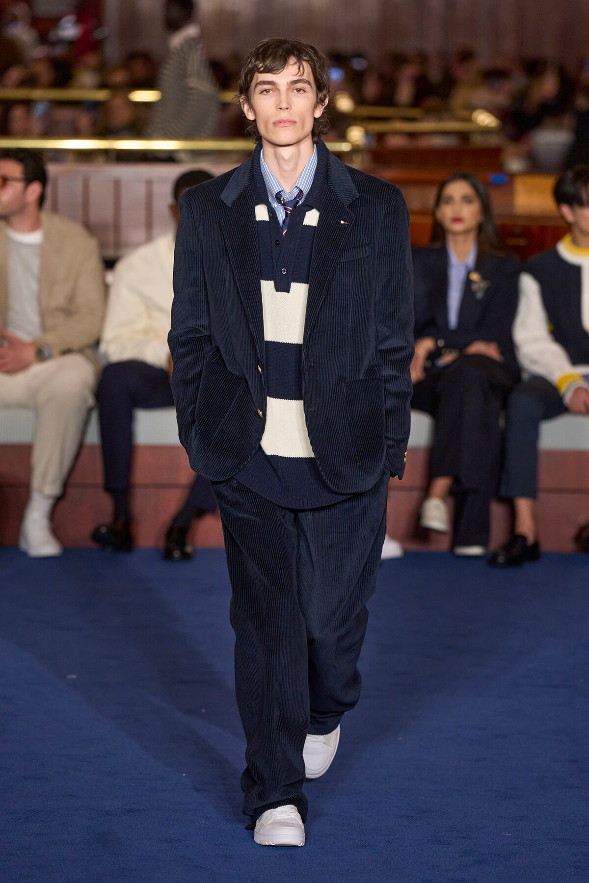 Male model walks down runway in navy corduroy suit with striped navy-and-cream sweater and tie at a fashion show. Audience in background.