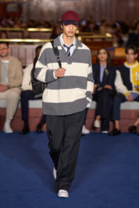 Male model walking a runway in a gray and white striped knit sweater, white sneakers, red cap, and a black backpack.