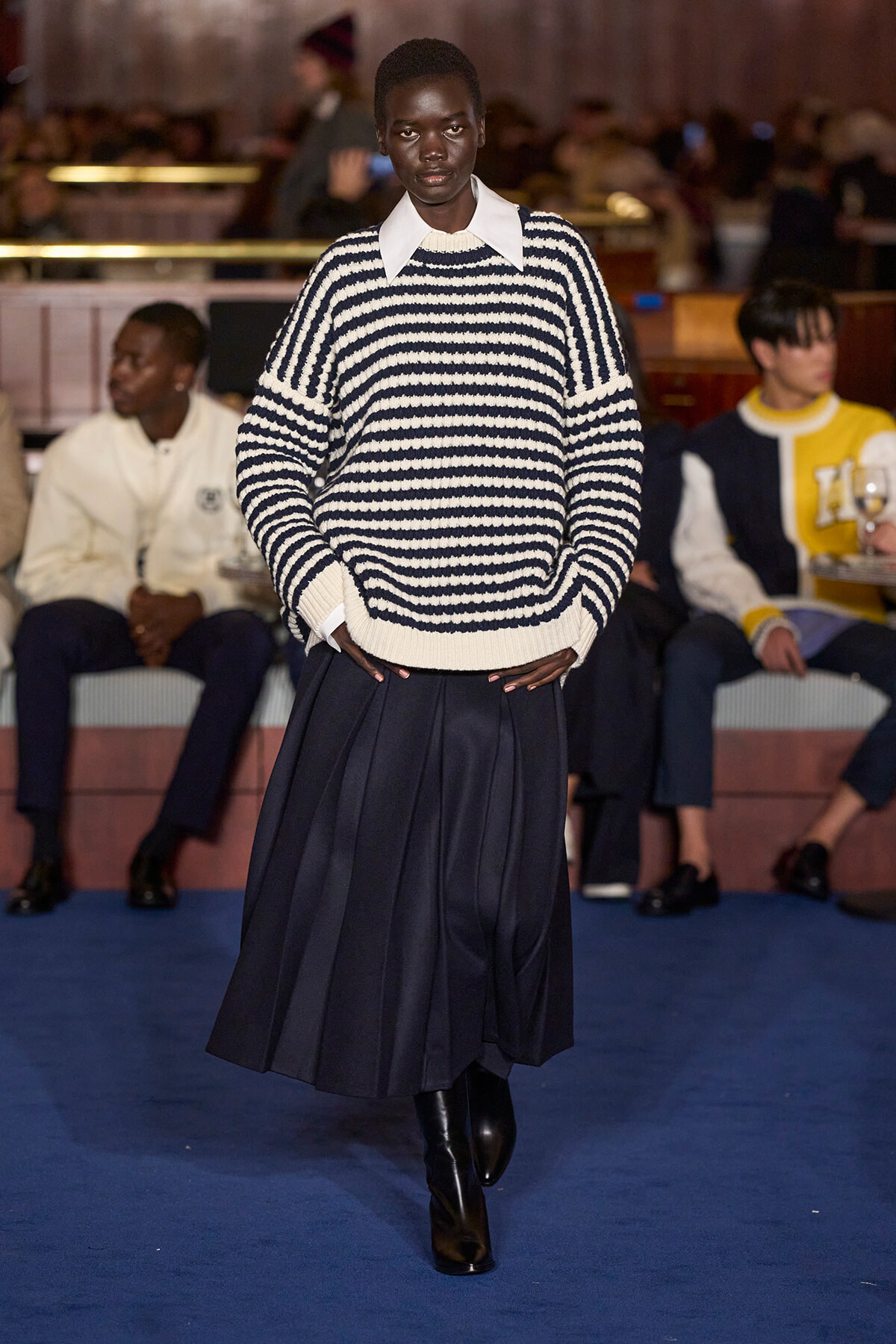 Model walks the runway in a navy-and-white striped knit sweater over a white collared shirt, paired with a pleated navy skirt; audience in background.
